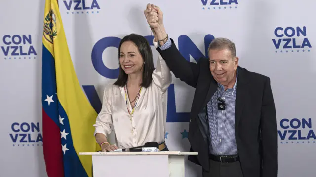 María Corina Machado y Edmundo González levantan sus brazos agarrados de la mano. En el fondo, una bandera de Venezuela y un logo que dice 