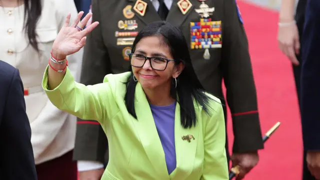 Delcy Rodríguez vistiendo una chaqueta amarillo limón saluda levantando la mano derecha.